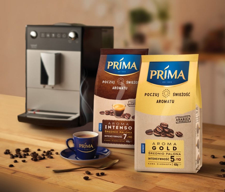 Produkty do kawy zbożowej Prima Aroma Gold i Prima Aroma Intenso z niebieską filiżanką kawy z logo Prima, na stole i ziarnami kawy przed ekspresem