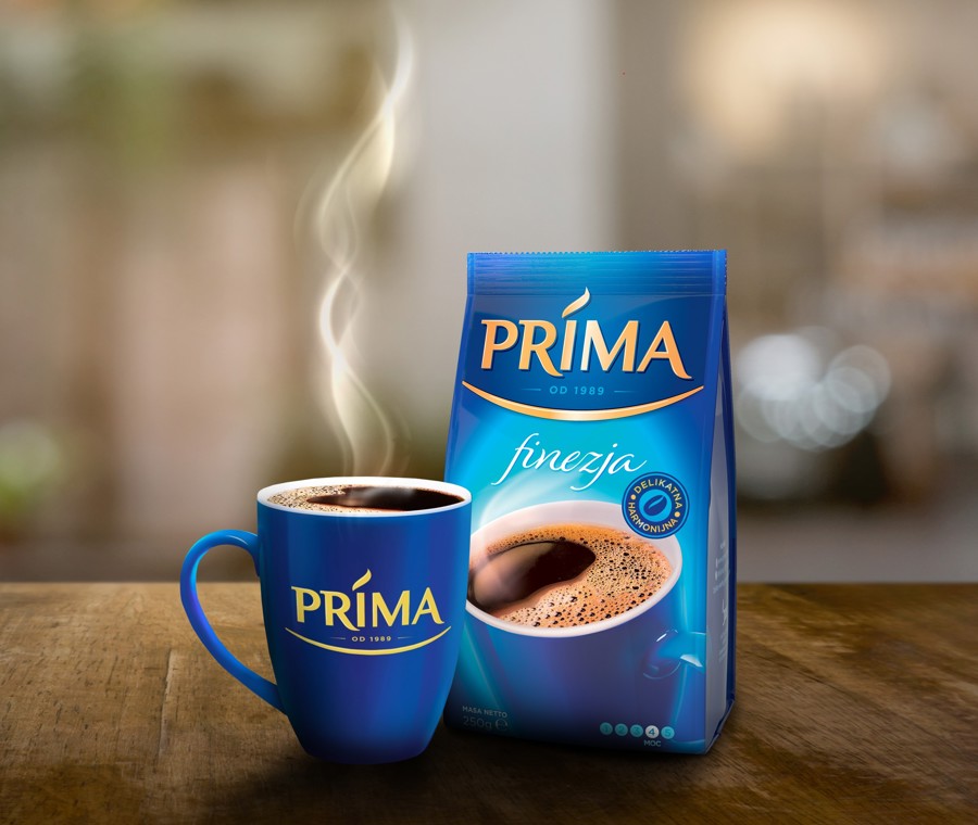 Opakowanie Prima Finezja z niebieską filiżanką kawy z logo Prima, na drewnianym stole i rozmytym tle
